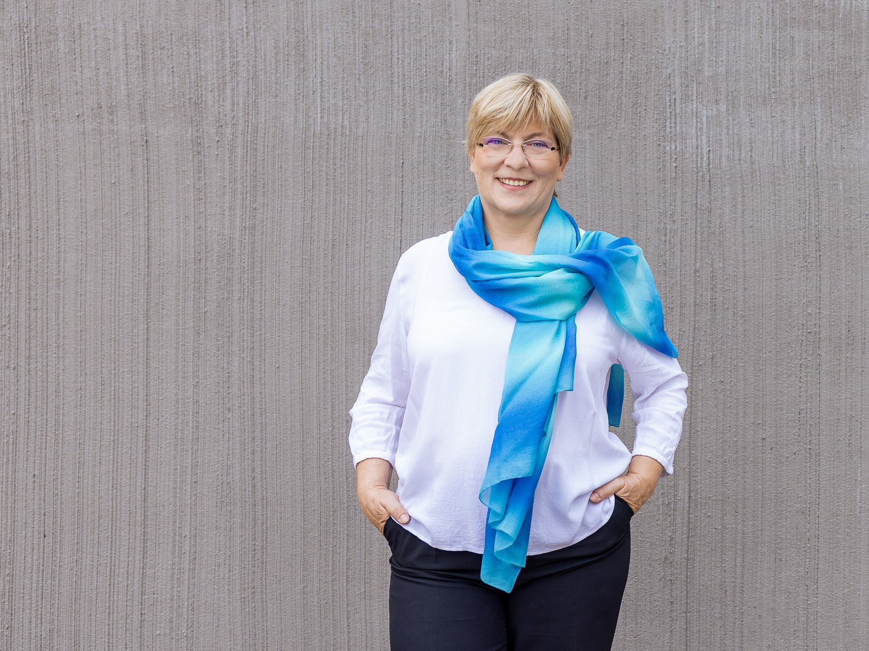 Frau mit kurzen Haaren steht vor einem Gebäude. Sie trägt eine weiße Bluse mit einem blauen Tuch. 