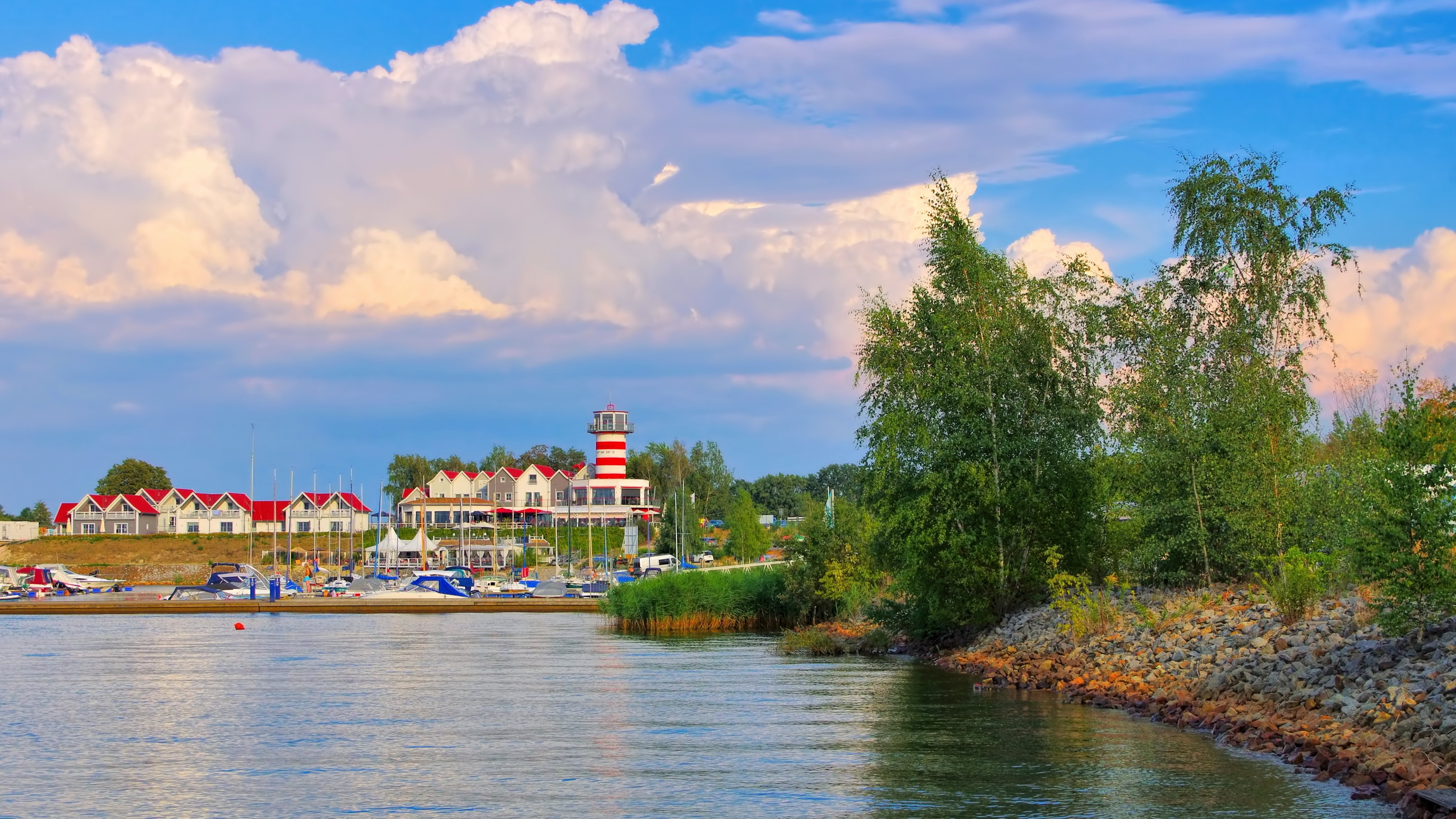 Bild: See mit bewachsenem Ufer, im Hintergrund Boote, Ferienhäuser und Leuchtturm