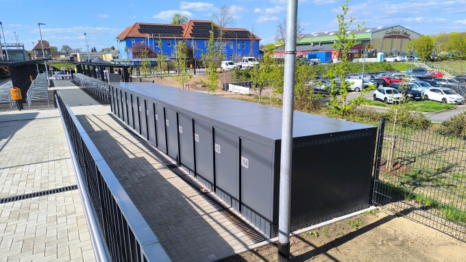 Boxen zum Einstellen von Fahrrädern, barrierefreier Zugang zur überdachten Fahrradabstellanlage am Bahnhof Erkner, rechts Parkplatz und Stadthalle Erkner, Mitte. blaues Gebäude mit Solardach